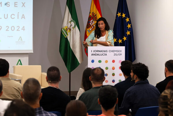 La consejera de Inclusión Social, Loles López, interviene durante las I Jornadas Chemsex de Andalucía el 25 de septiembre de 2024. Al fondo se observan las banderas de Andalucía, España y la Unión Europea, mientras ella se dirige a un grupo de asistentes en una sesión formativa sobre chemsex, organizada por la Junta de Andalucía